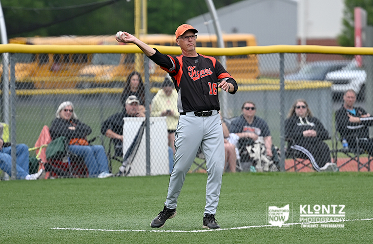 Baseball pitcher in black and orange uniform mid-throw on the mound with spectators behind a chain-link fence.