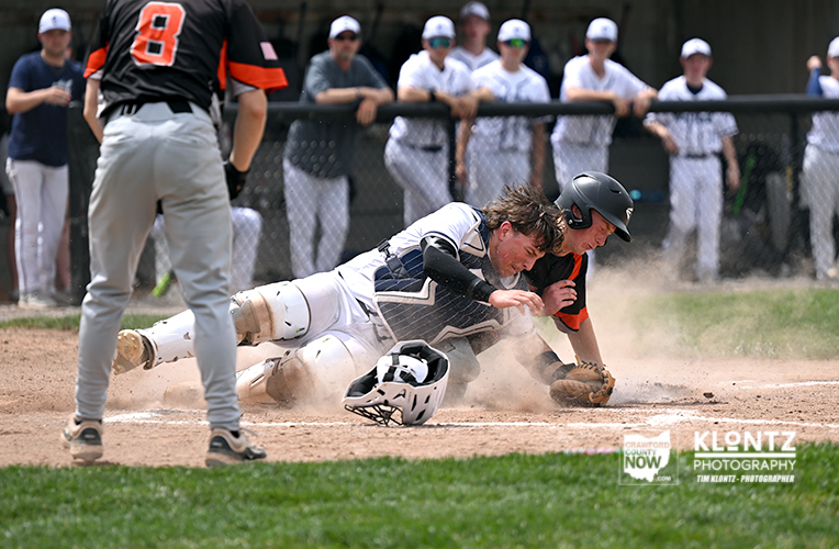 Baseball catcher and sliding runner collide at home plate, dust kicking up as spectators watch from the fence.