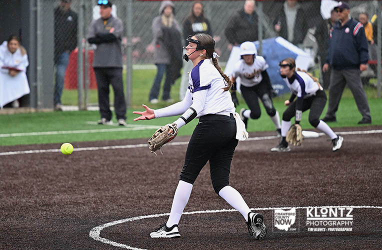 Female softball pitcher in white and purple uniform releases a pitch on a dirt infield with teammates and spectators in the background.