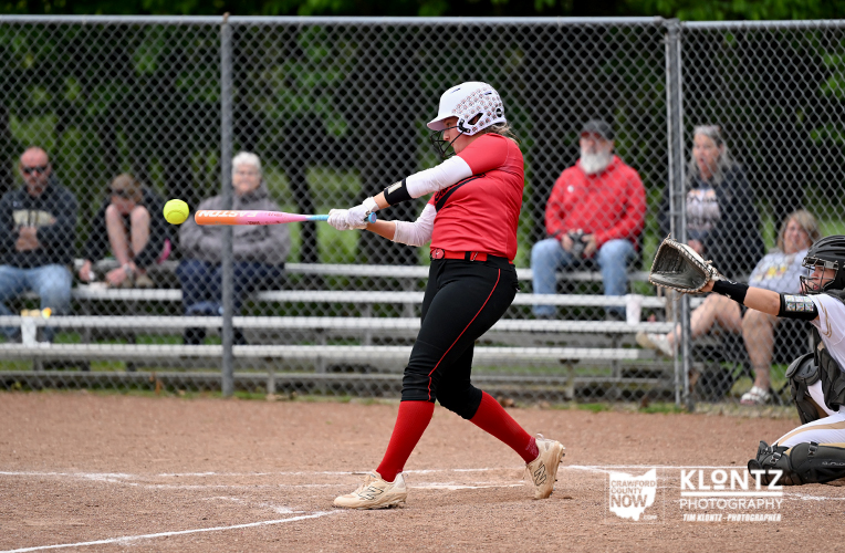 Female softball batter in red jersey swings at a pitched ball on a dirt field; catcher ready behind home plate; spectators behind chain-link fence.