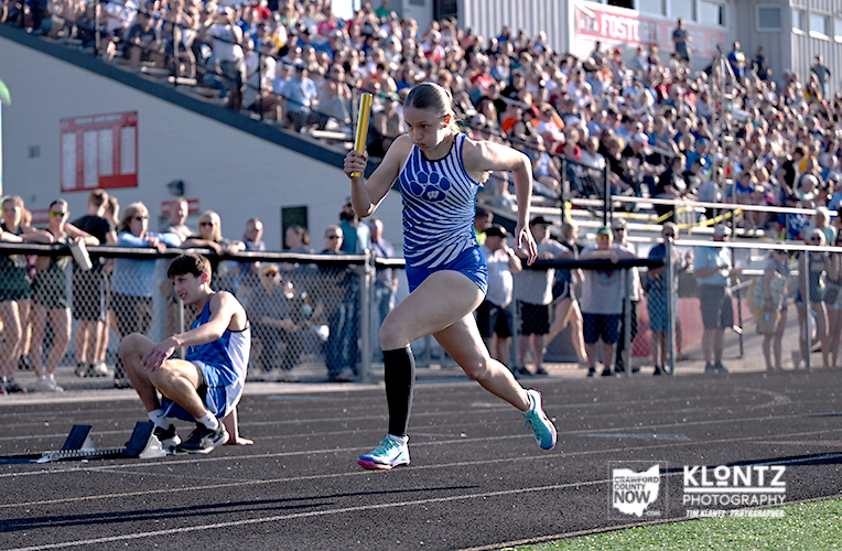 Female relay runner in a blue striped uniform sprints with a baton on a track as a crowd watches from the stands.