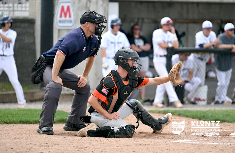 Baseball catcher in black and orange gear reaches with his glove at home plate while an umpire crouches behind him on the dirt field