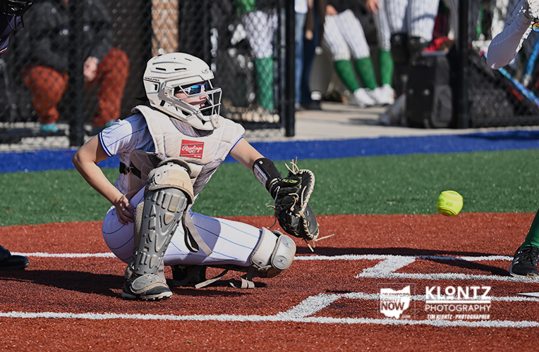 Youth baseball catcher in full gear crouches on home plate, glove ready as a bright yellow ball sails nearby.
