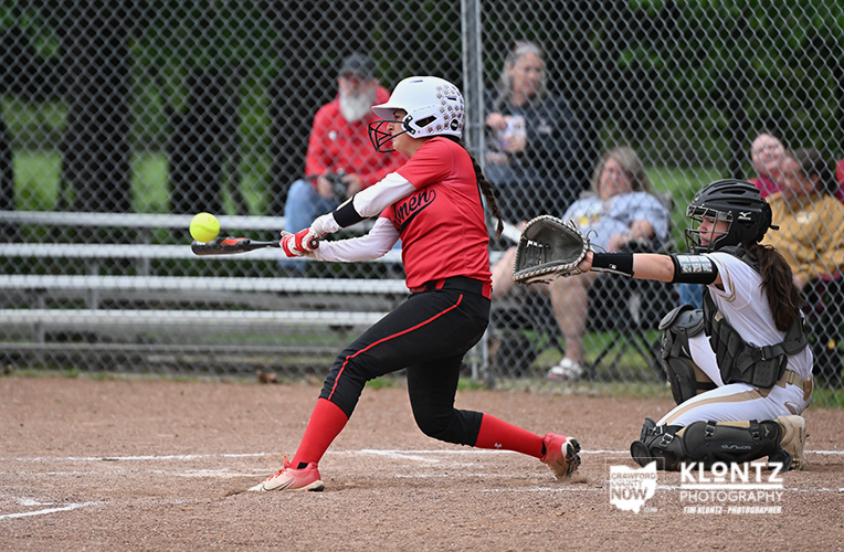 Female softball batter in a red uniform swings at a pitch, with the catcher ready behind home plate and spectators in the background.