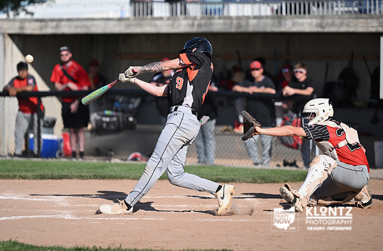 Baseball batter in black and orange swings at a pitch as the catcher in red and white gear crouches behind home plate.