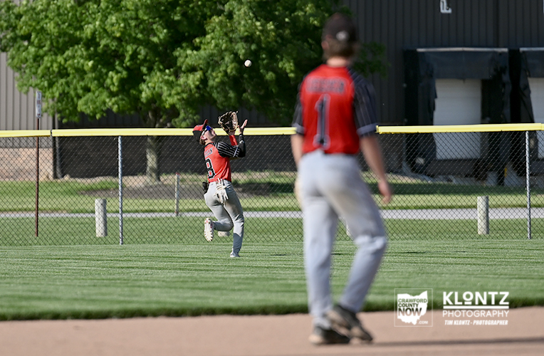 Youth baseball game action: a red-jersey fielder reaches up with his glove to catch a ball near the outfield fence, another player blurred in foreground.