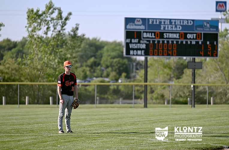 A young baseball fielder in a black and orange uniform stands on a grassy field near a scoreboard at Fifth Third Field, glove in hand.