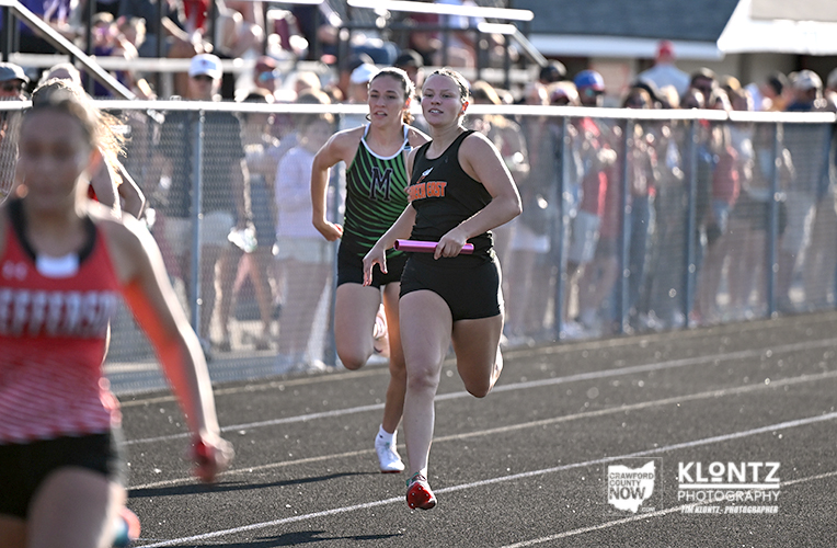 Two female relay runners sprint on an outdoor track while a crowd watches from behind a chain-link fence; one carries a pink baton.