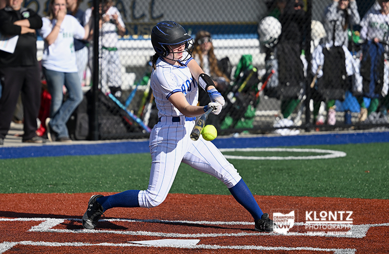 Female softball player in a white pinstripe uniform swings at a pitched ball during a game on a green and red field, spectators behind a chain-link fence.