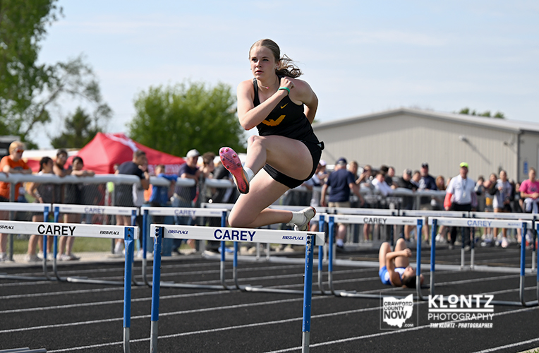Female hurdler in a black uniform clear a hurdle during an outdoor track race, with spectators in the background.