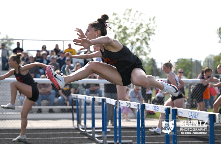 Female hurdler in a black uniform clears a hurdle on a track during a race, with other competitors and spectators in the background.