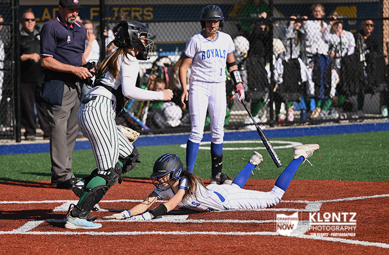 Runner slides headfirst into a base as the catcher and umpire anticipate the play on a softball field at the edge of the dirt and turf.