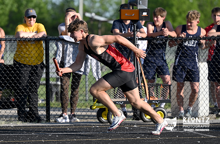 Male sprinter in red-and-black uniform launches from the starting line, baton in hand, as spectators watch behind a chain-link fence.