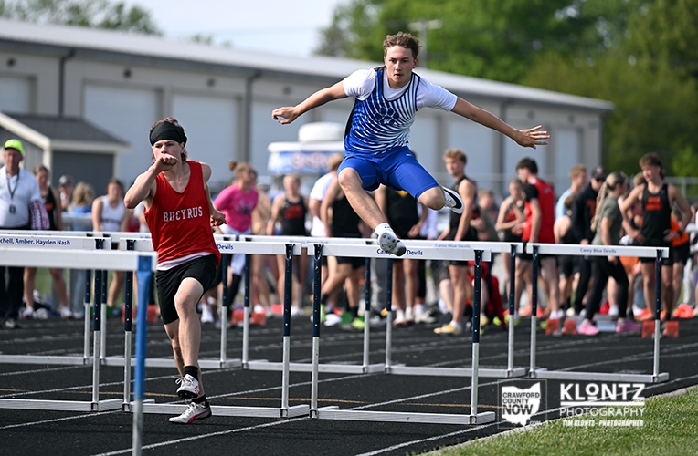 Male hurdler in blue and white clears a hurdle while a rival in red runs toward the next barrier, with spectators in the background on a track.
