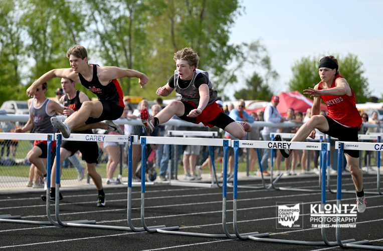 Three male hurdlers leap over hurdles in a daytime track race, with spectators behind them on the field.
