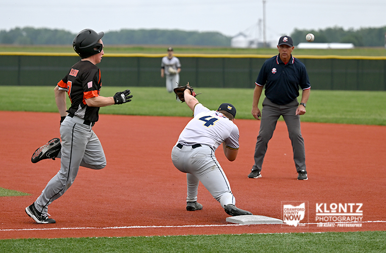 Baseball game action: a runner slides into a base as a fielder approaches, while an umpire watches from the dirt field.