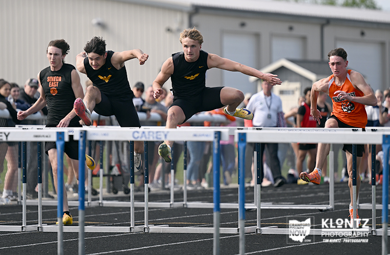 Four male sprinters leap over hurdles in a track and field race, with spectators and a building in the background.