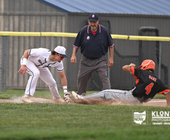 Baseball play at second base: fielder in white reaches for the bag as a runner in black slides, ball in the air and dust around the base, with an umpire watching.