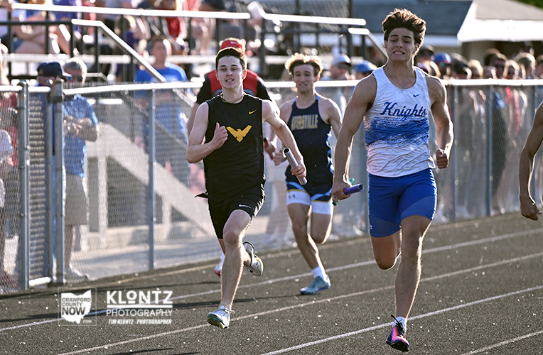 Track runners in a relay race sprinting for the baton on a crowded stadium track.