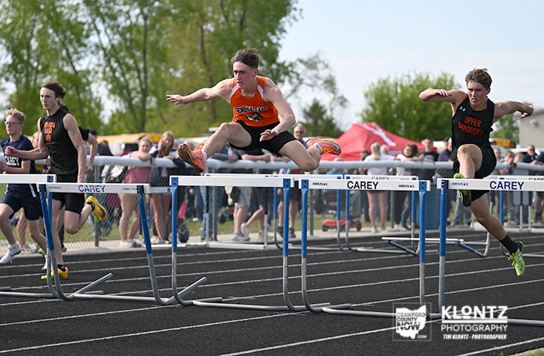 Male hurdlers jump over blue-and-white hurdles on a track; central runner in orange jersey is mid-air during a race.