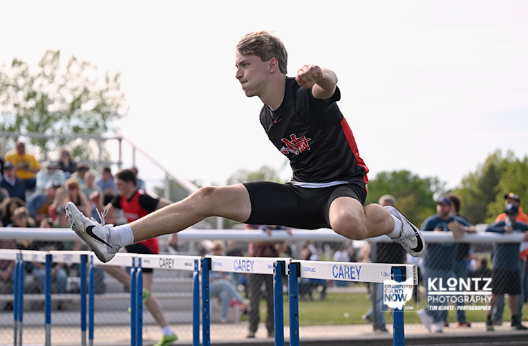 Male athlete in a black uniform jumps over a hurdle at an outdoor track meet, with spectators in the background.