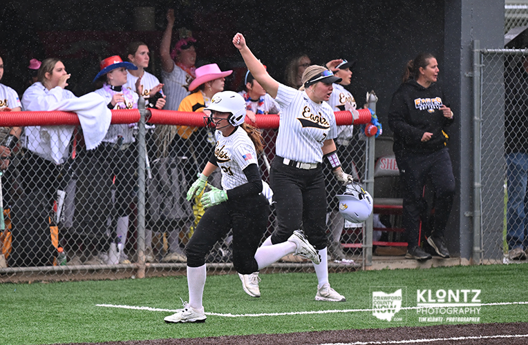 Two softball players in white uniforms run along the baseline after a play, with cheering fans behind a chain-link fence.