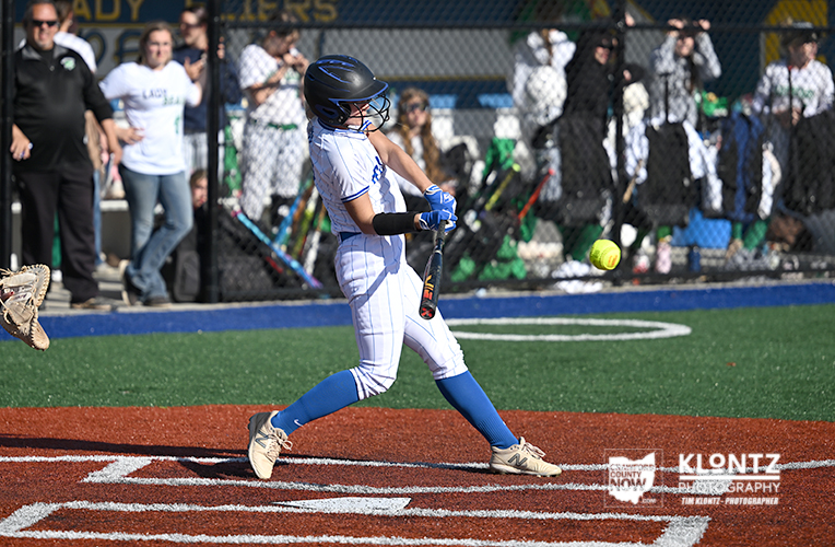 Young baseball player in a white uniform swings at a pitch near home plate with spectators behind a chain-link fence.