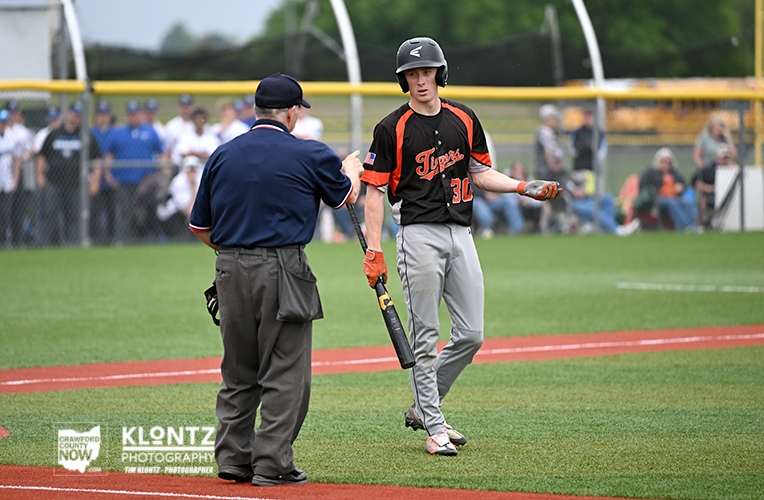 Baseball batter in black and orange uniform high-fiving an umpire at home plate during a game, with spectators in the background.