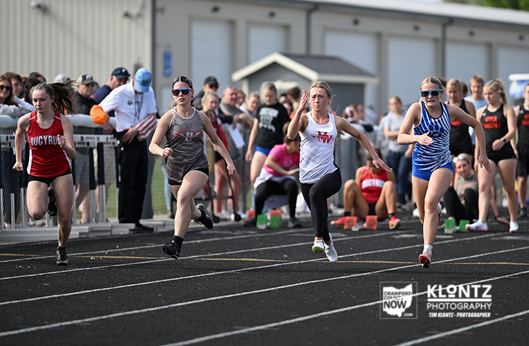 Group of young female sprinters racing on a track with spectators in the background.