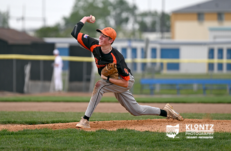 Young baseball pitcher in an orange cap and black jersey throws a pitch from the mound during a game on a grassy field.