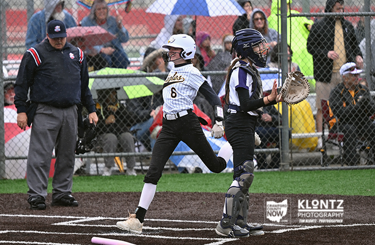 Young female softball player in a white striped jersey and helmet runs toward home plate as the catcher in dark gear prepares nearby, with spectators behind a chain-link fence.