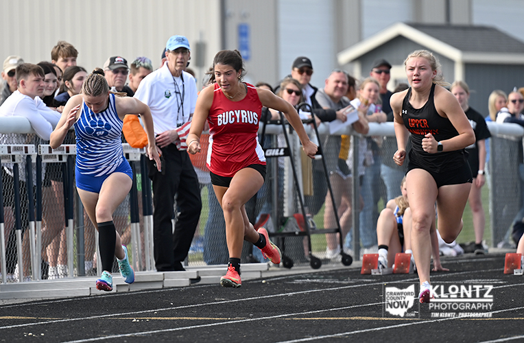 Three female sprinters race on a track with a cheering crowd behind a fence, red center runner wearing Bucyrus and blue/black opponents flanking her.