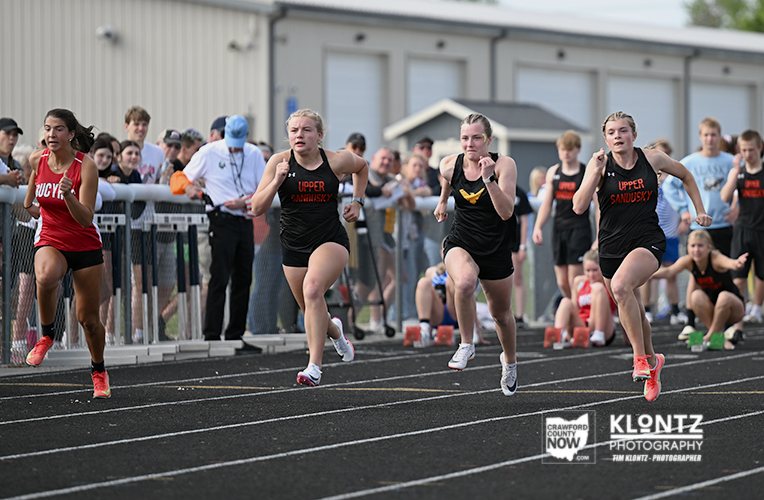 Four female sprinters in black Upper Sandusky uniforms lead a track race on an outdoor track, with a red-clad runner on the left and a crowd watching behind the fence.