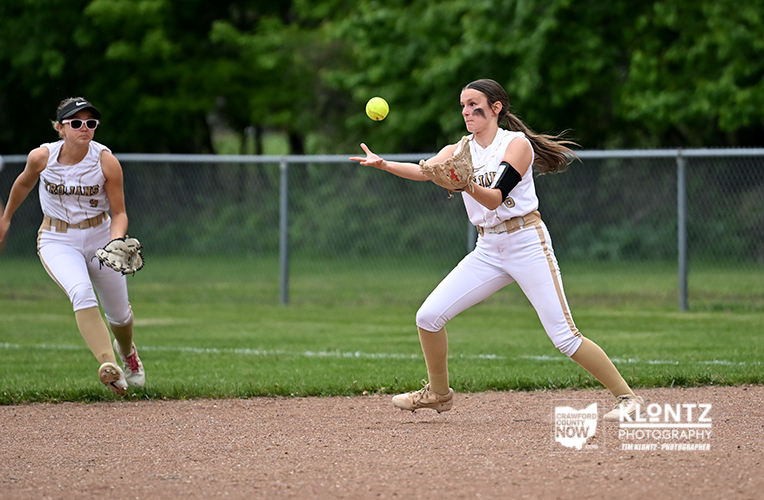 Two female softball players in white uniforms on a dirt infield, one in the foreground reaching with her glove as a ball flies toward her while a teammate nears in the background.