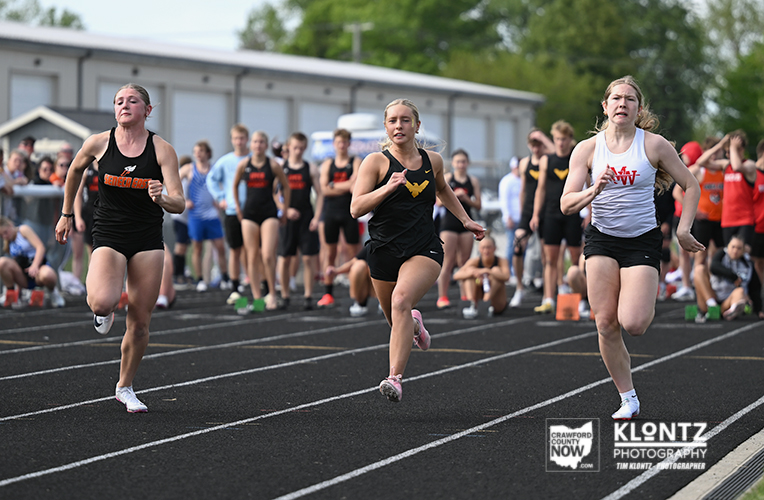 Three female sprinters race down a track with a crowd in the blurred background. A black-uniform runner is on the left, a center black-uniform runner with a yellow emblem, and a white-top runner on the right.