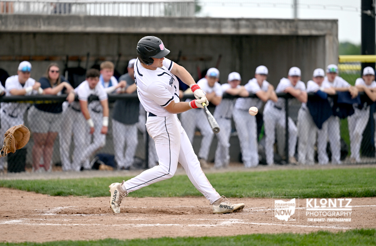 Baseball batter in white uniform swinging at a pitch, helmet on, on a dirt infield with dugout and spectators in the background.
