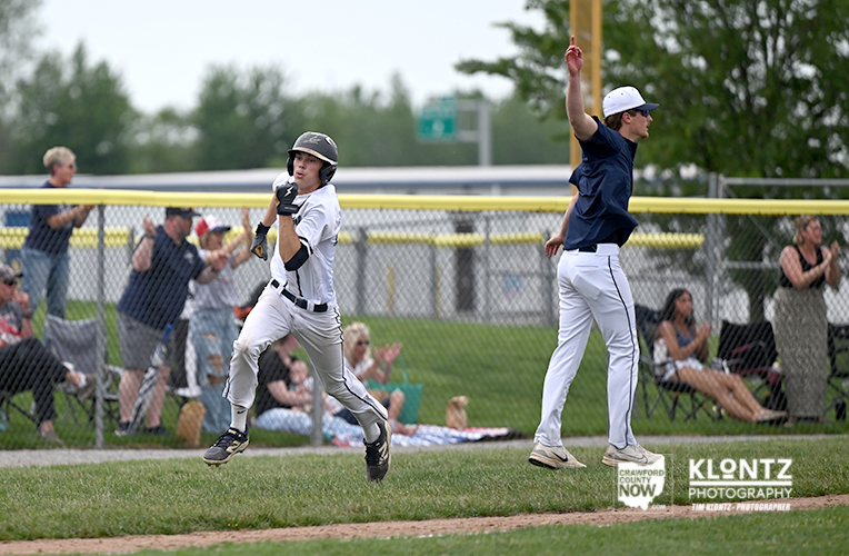 Youth baseball game action: a runner in a white uniform and helmet sprinting toward home plate while a batter in a navy shirt swings nearby; spectators sit behind a chain-link fence in the background.