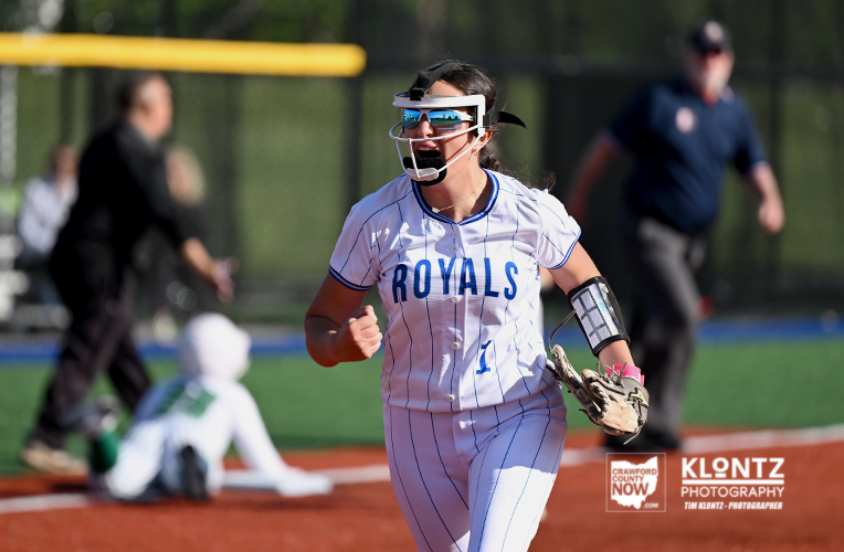 Softball player in a white Royals uniform runs on the field, wearing a protective face mask, glove on her left hand, celebrating a play.