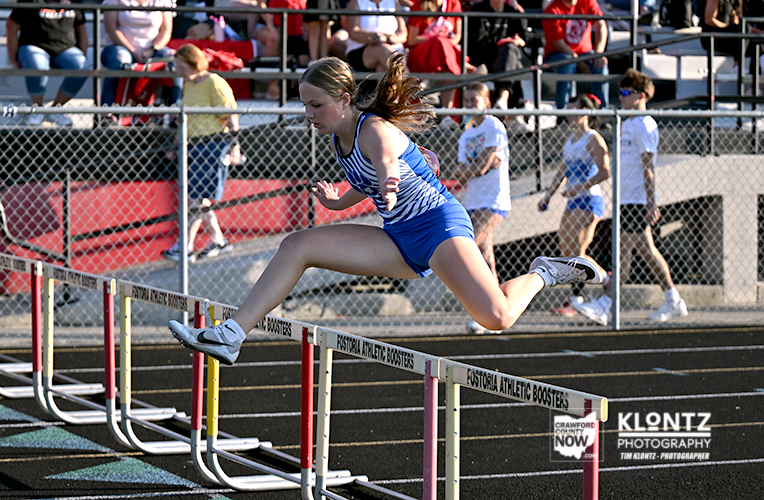 Female hurdler in a blue uniform clears a hurdle during an outdoor track race, with spectators in the stands behind her.