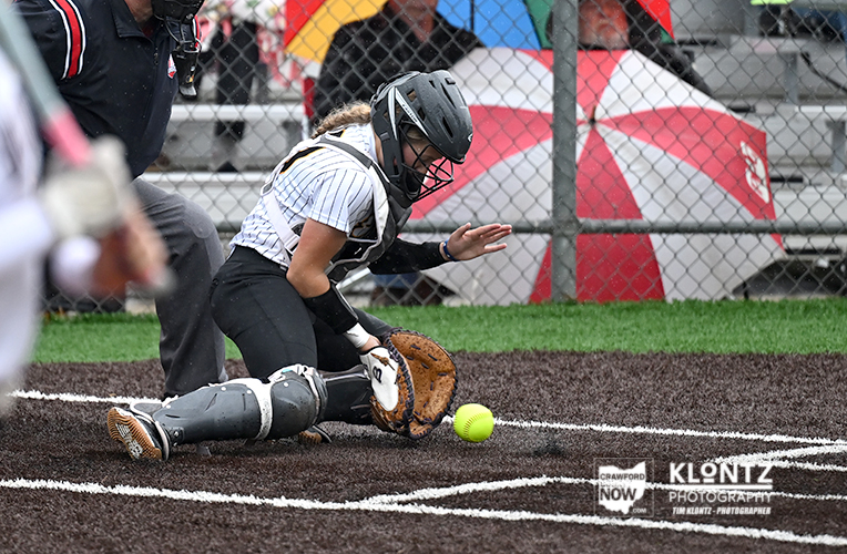 Softball catcher in gear dives to stop a ground ball near home plate, glove low to the turf, with a bright yellow ball nearby.