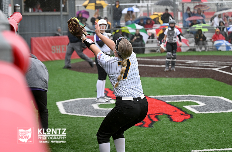 Female softball pitcher in a striped jersey mid-throw on a wet field as spectators under umbrellas watch from the stands in the rain.