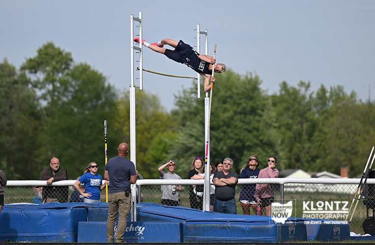 Pole vaulter in mid-air clearing a horizontal bar between two vertical poles as spectators watch from behind a fence.