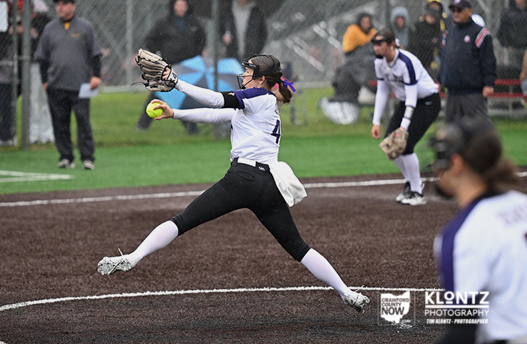 Female softball pitcher in white and purple delivering a pitch on a dirt mound with spectators watching behind a chain-link fence.