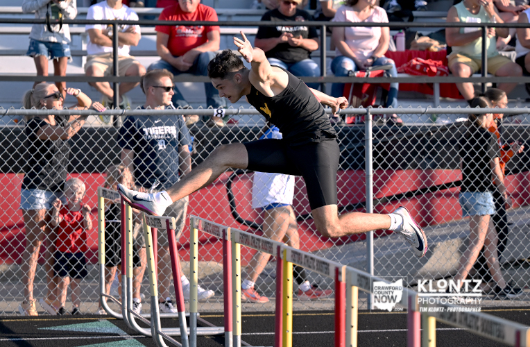 Male hurdler in a black uniform jumps over a hurdle on an outdoor track, with spectators behind a chain-link fence.