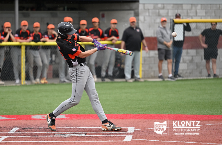 Youth baseball batter in black and orange uniform swings a colorful bat at a pitched ball during a game, with teammates and spectators behind a chain-link fence.