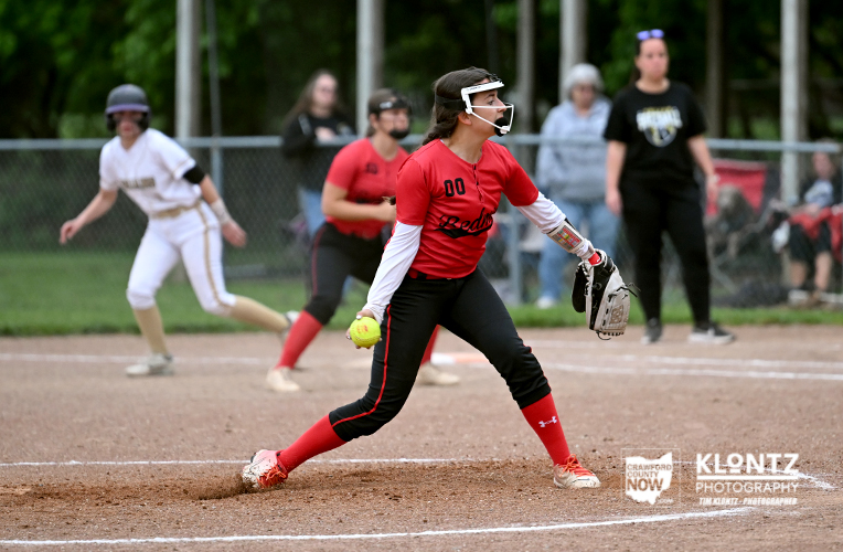 Female softball pitcher in a red jersey winds up on a dirt field with a glove and helmet, teammates and spectators in the background.