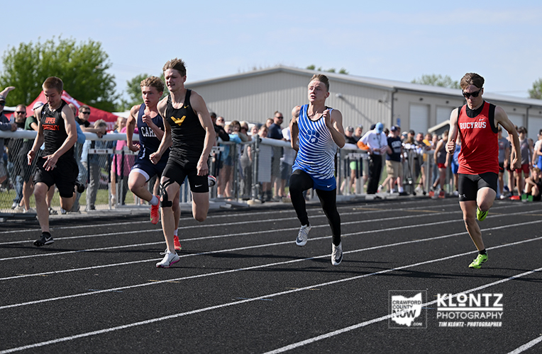 Five male sprinters race on a track, with a cheering crowd behind a fence on a sunny day.