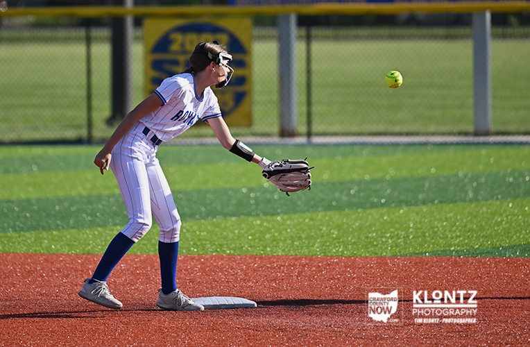 Softball player in a white uniform with blue socks fields a ball near first base on a sunny field.