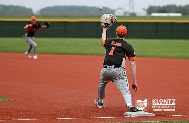 Baseball pitcher in a black and orange jersey (#1) throws a pitch on a red clay infield; a teammate watches from the background.
