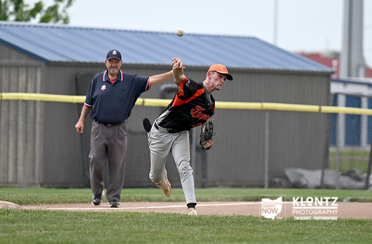 Baseball pitcher in orange cap delivering a pitch as a ball sails through the air, with an umpire standing behind on the infield.